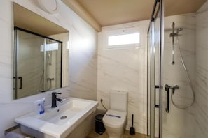 Casa de banho com lavatório branco, espelho, sanita e chuveiro com porta de vidro, e janela.