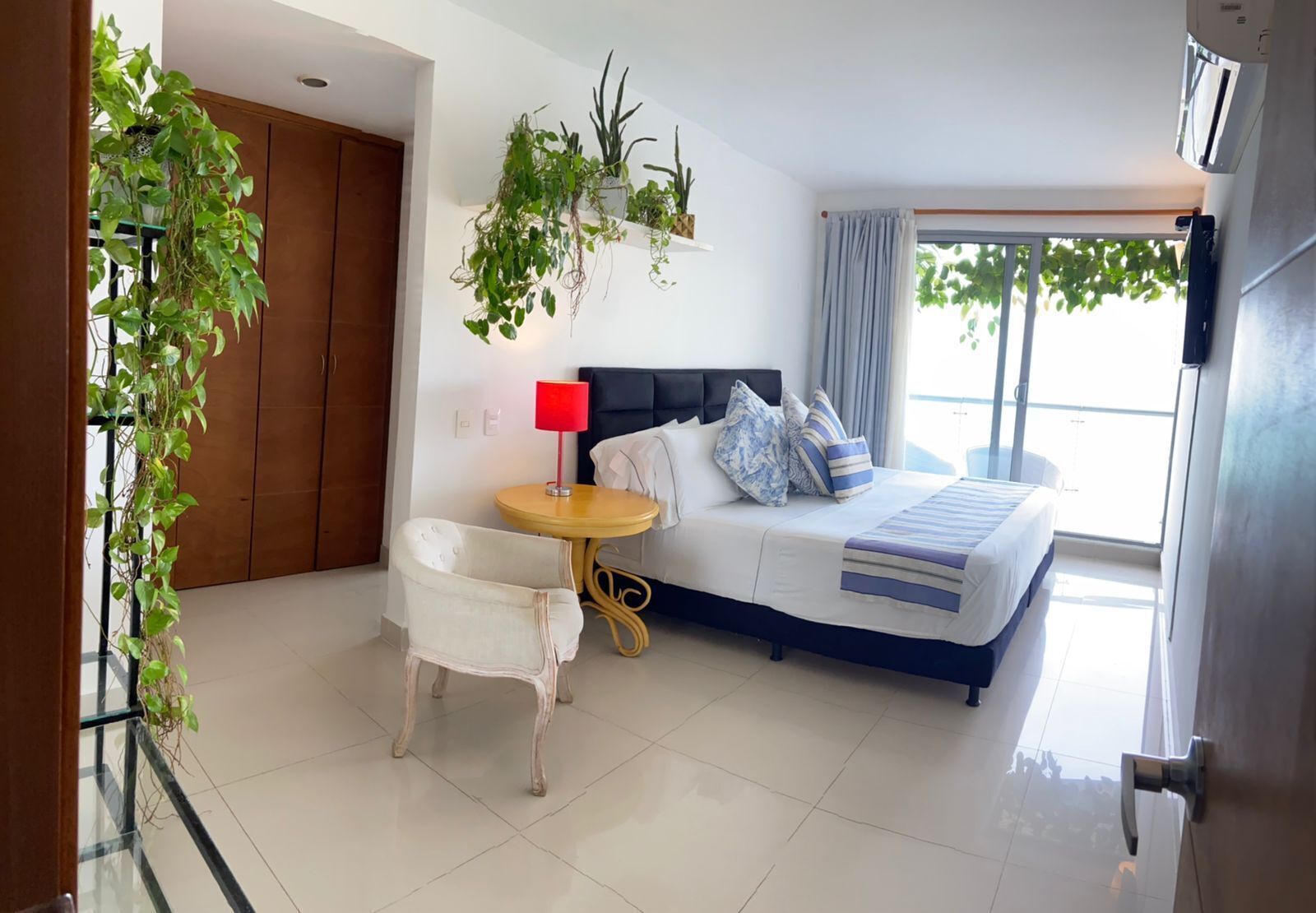 Una habitación de hotel moderna con una cama, una mesita de noche con lámpara, una silla y plantas en macetas.
