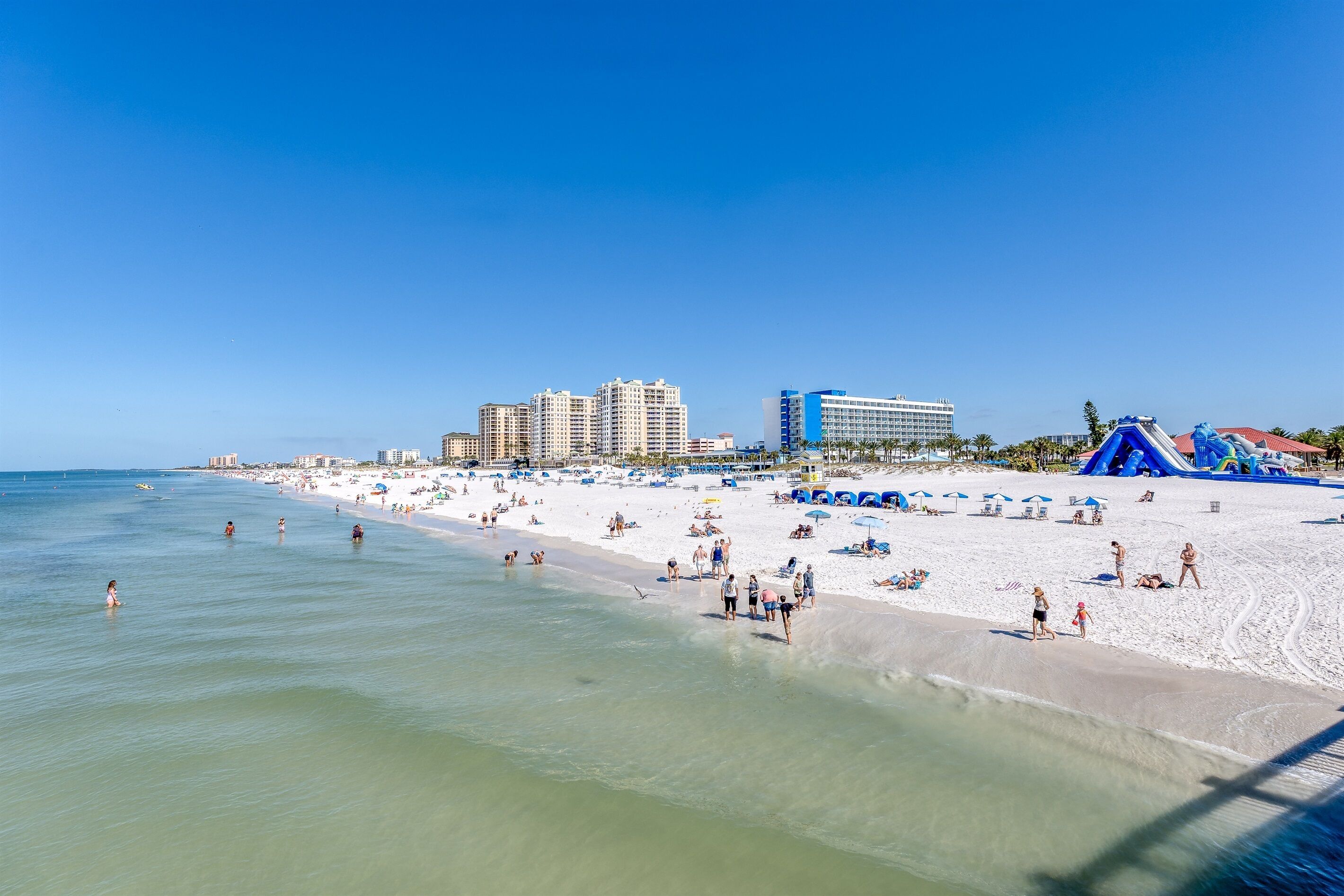 Una playa con arena, agua cristalina y varios edificios al fondo.