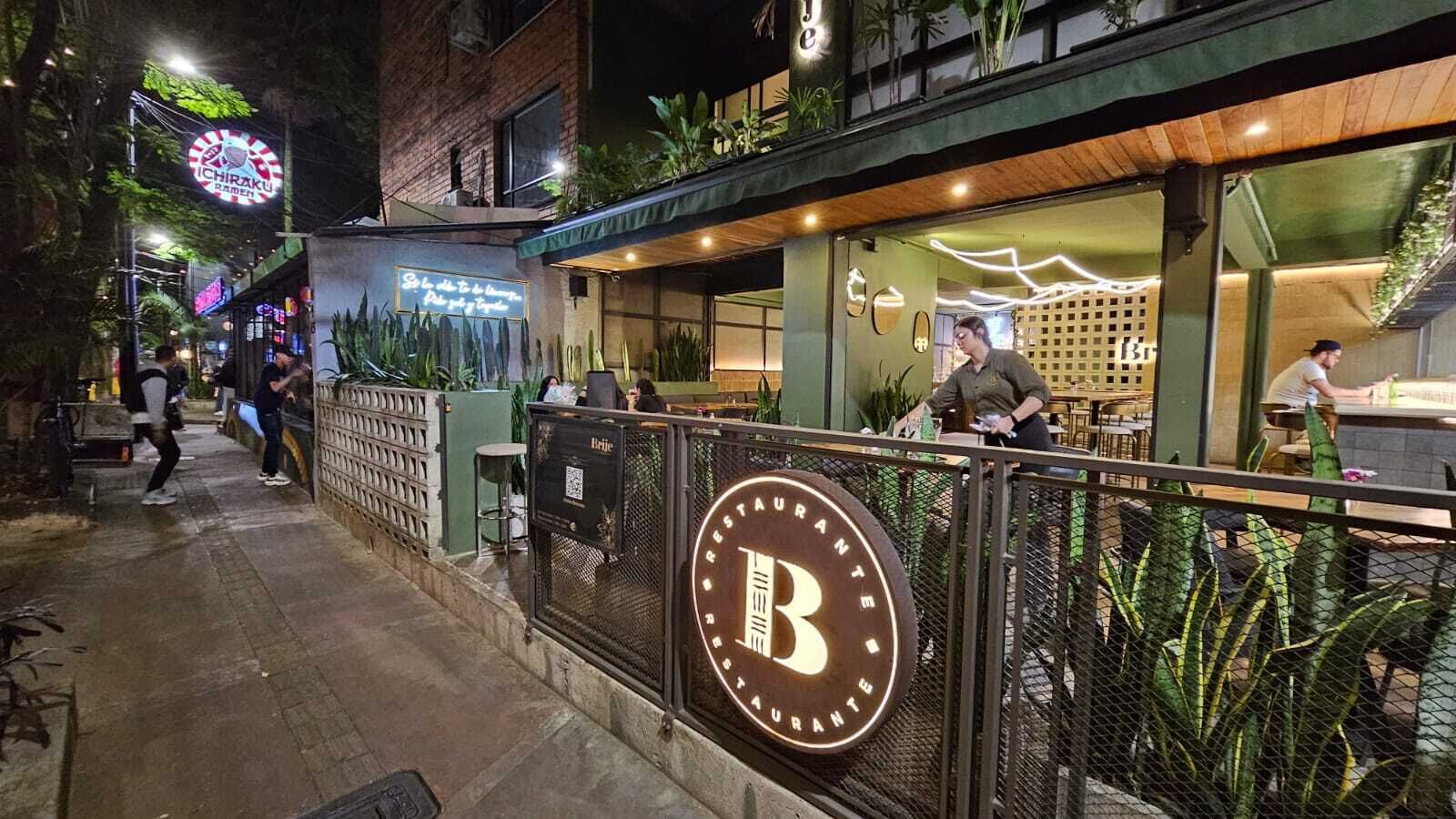 A restaurant with a prominent illuminated 'B' sign, outdoor seating, and a neon sign in the background.