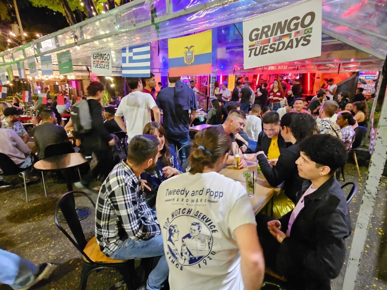 A crowded outdoor dining area with tables and chairs, under a transparent roof structure.