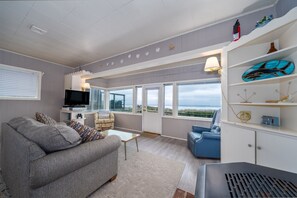 A modern living room with a grey sofa, a blue armchair, a TV, and a large window offering a view of the sea. - Steps to the Beach, Charming Oceanfront Bungalow with Deck and Ocean Views (Rockaway Beach)