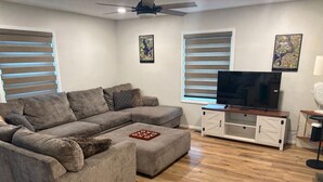 Uma sala de estar com sofá seccional, uma TV de tela plana sobre um suporte de madeira e duas janelas com persianas.