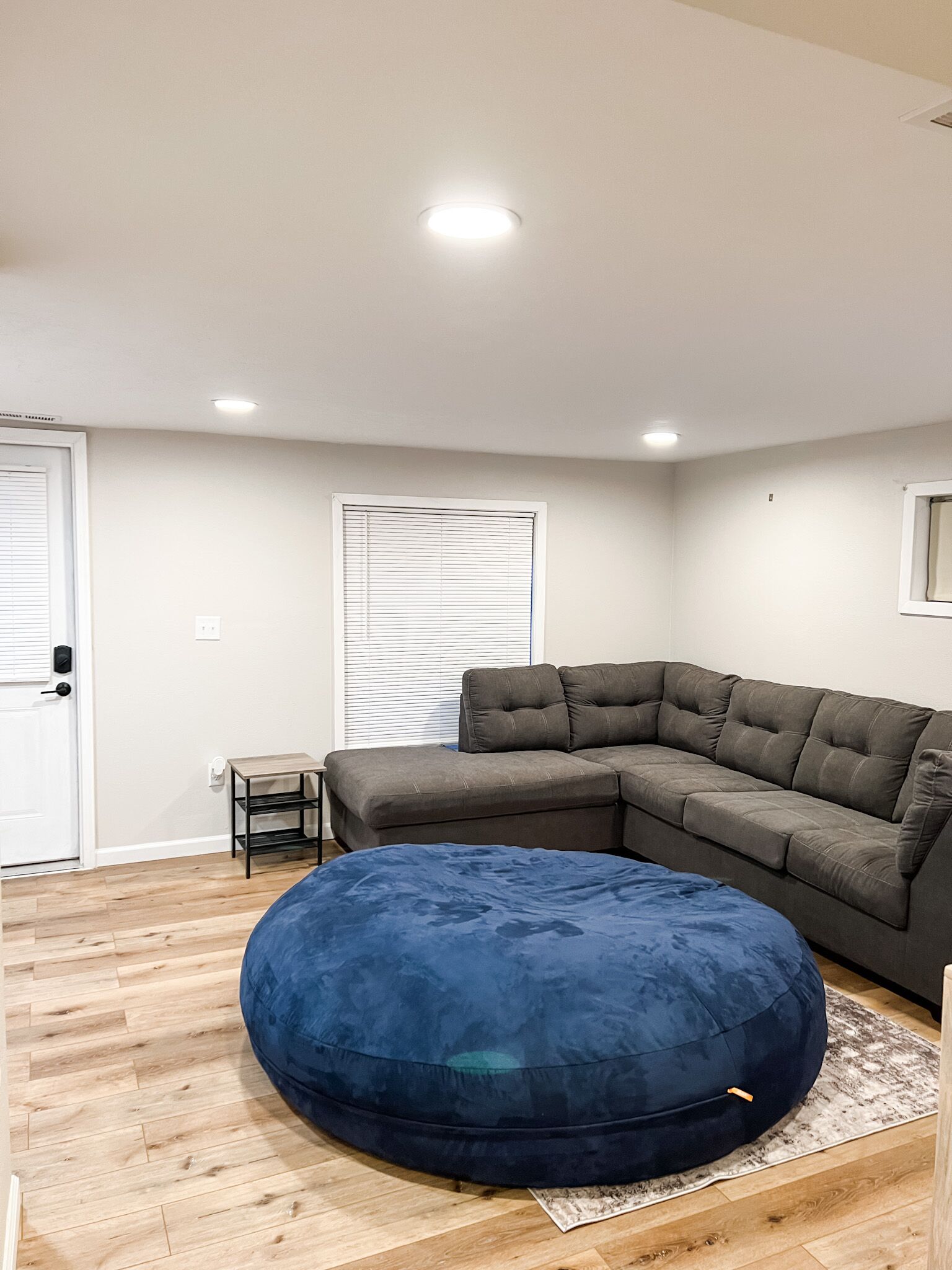 A living room with a sectional sofa, a round bean bag, a small table, and a window with blinds.