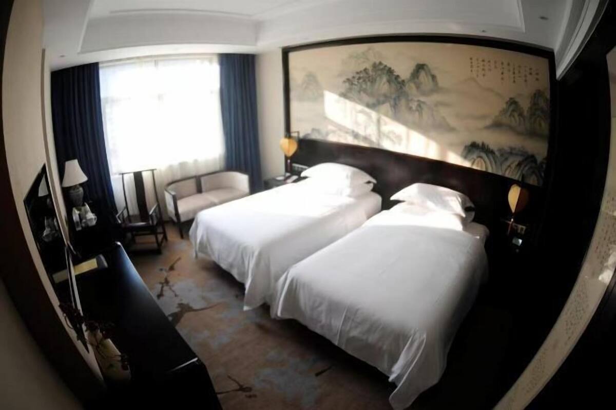 A hotel room with two beds, a desk, a chair, a painting on the wall, and a window with curtains.