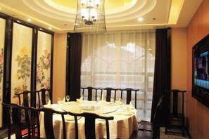 A dining area with a round table set for four, surrounded by wooden chairs, with a traditional Chinese screen and a large window with curtains. - Baodeli Hotel (Pan'an)
