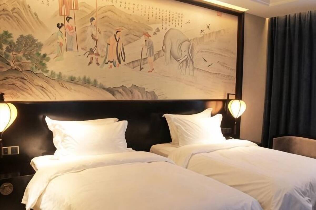 A hotel room with two beds, a large traditional painting on the wall, and a nightstand with a lamp.