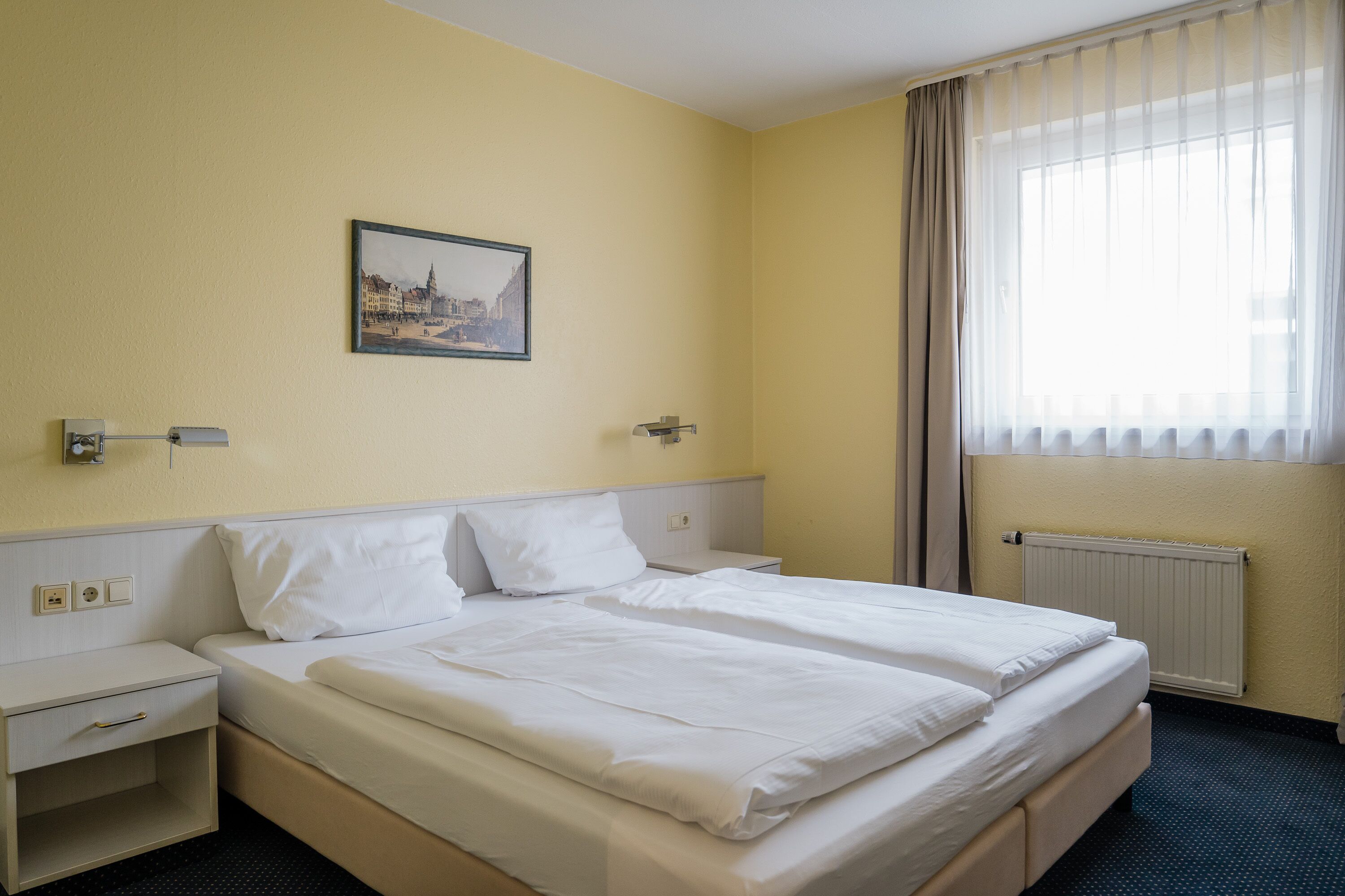 A hotel room with a double bed, a nightstand, a wall-mounted picture, and a window with curtains.