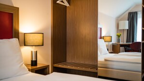 A hotel room with a bed, a nightstand with a lamp, a wooden wardrobe, a desk with a chair, and a window with curtains. - Point Hotel Apartments & Rooms (Dresden)