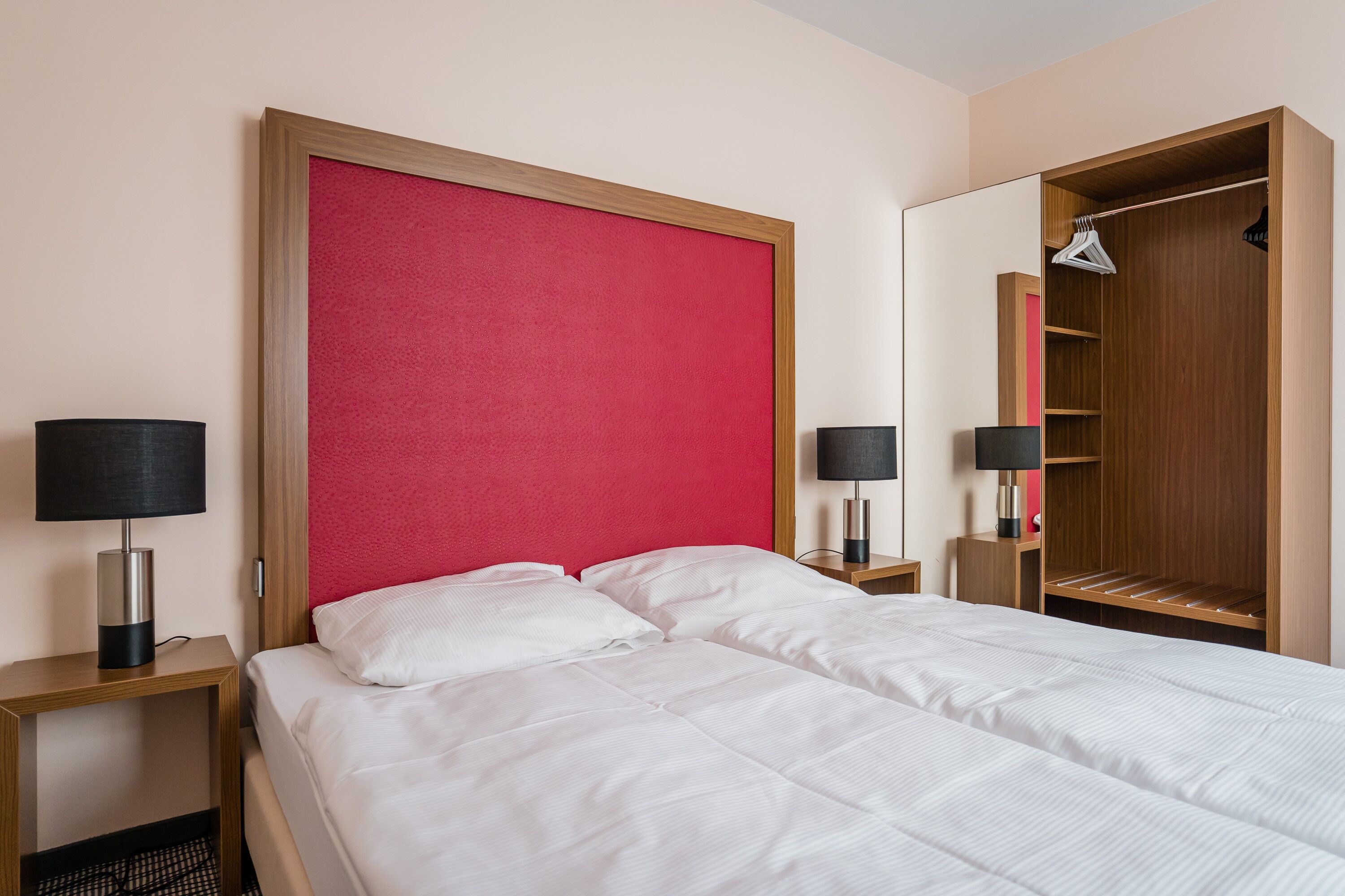 A hotel room with a bed, two bedside tables, a red headboard, a wardrobe, and a mirror.