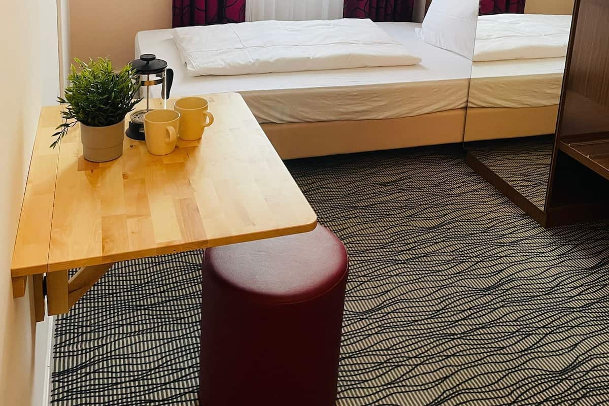 A hotel room with a bed, a small table with a plant and two cups, a red ottoman, and a patterned carpet.