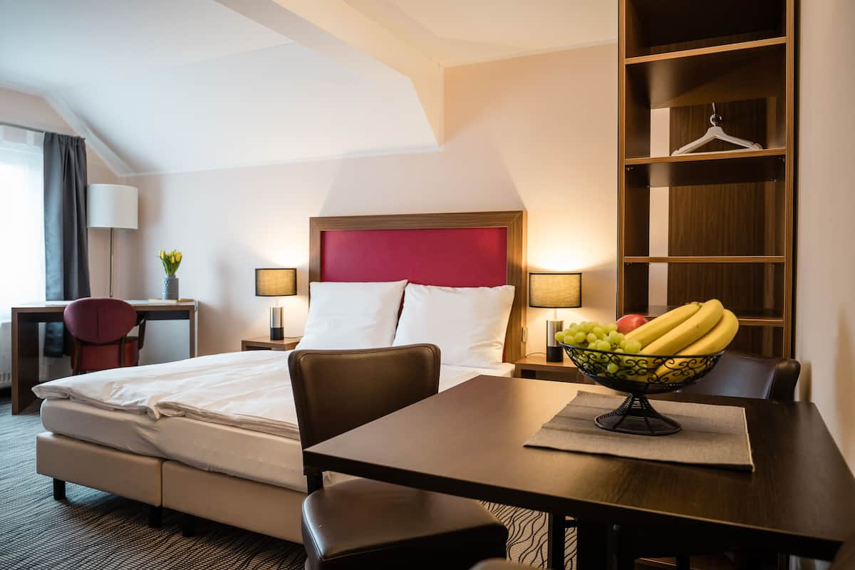 A hotel room with a bed, bedside table, lamp, chair, and a dining table with a fruit bowl.