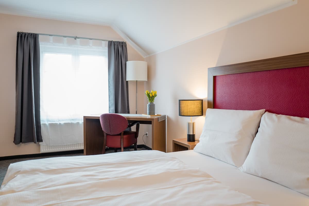 A hotel room with a large bed, a desk, a chair, a lamp, and a window with curtains.
