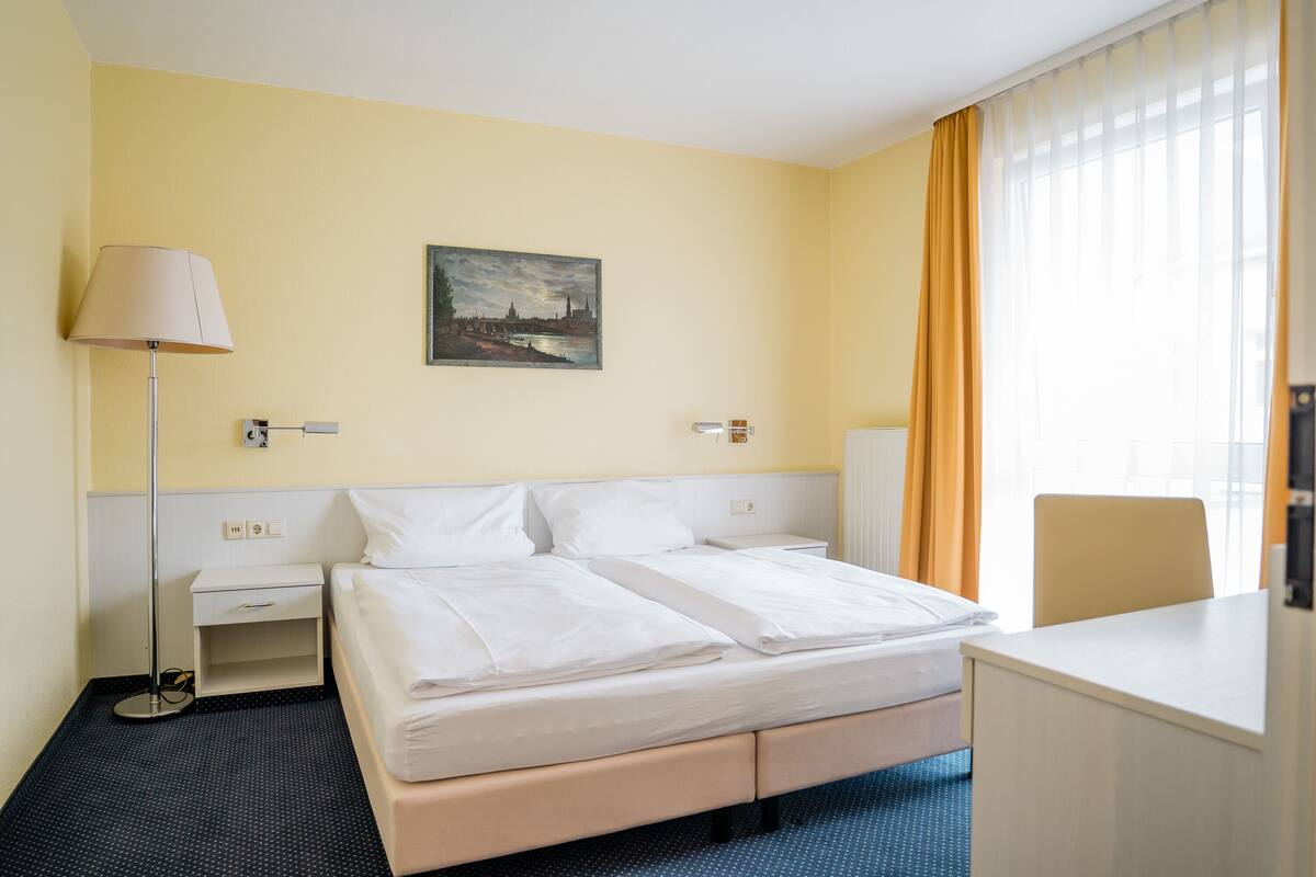 A hotel room with a bed, bedside table, lamp, desk, chair, and a painting on the wall.