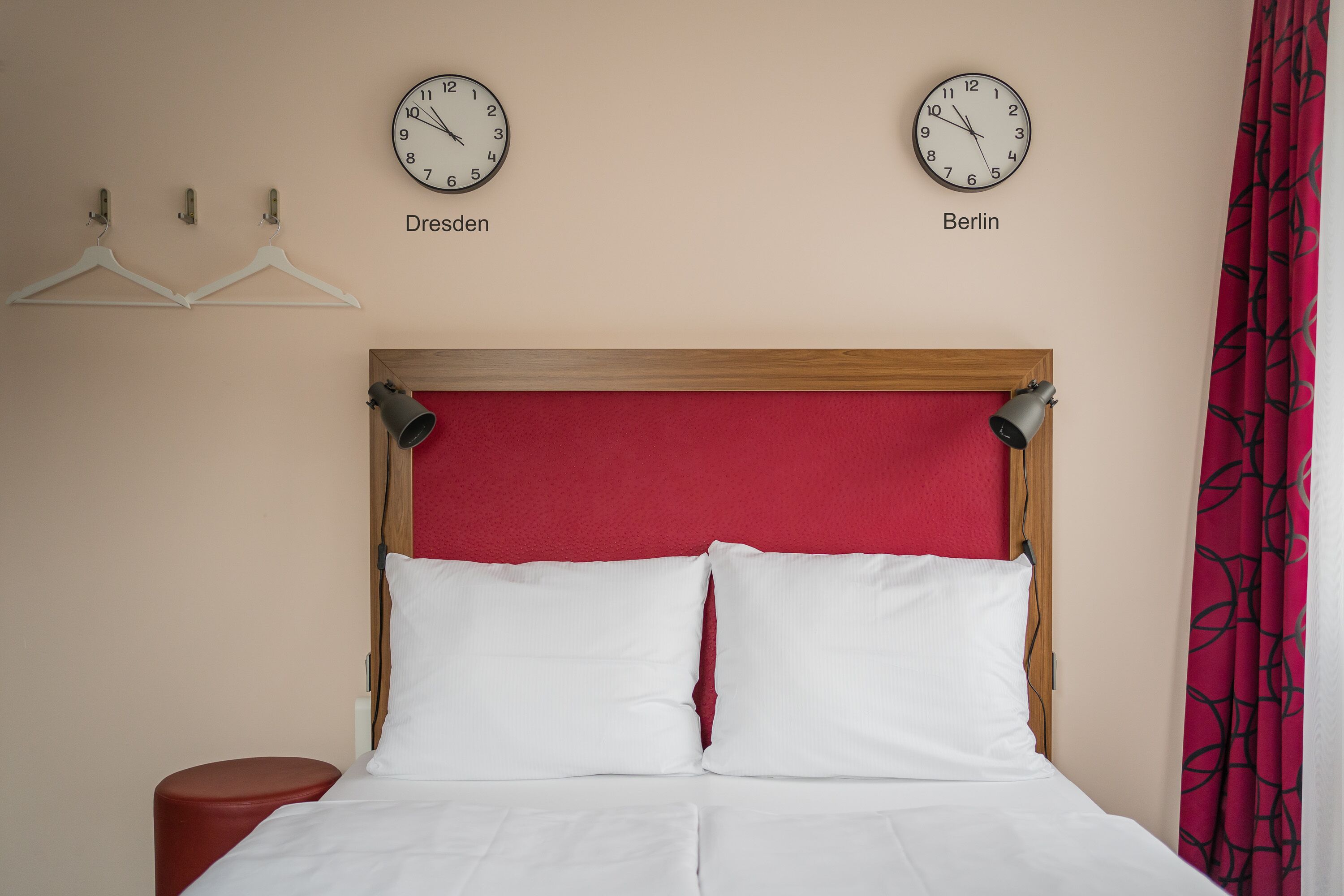 A hotel room with a bed, two clocks labeled 'Dresden' and 'Berlin', and a red curtain.