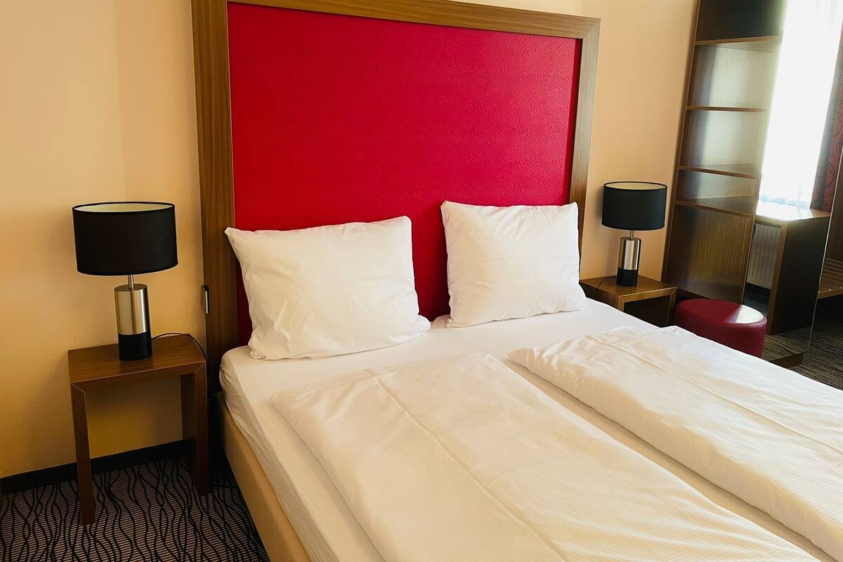 A hotel room with a large bed, two bedside tables with lamps, a red accent wall, and a mirror.