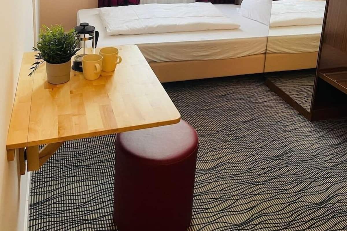 A hotel room with a bed, a desk with a plant and mugs, a red stool, and a patterned carpet.