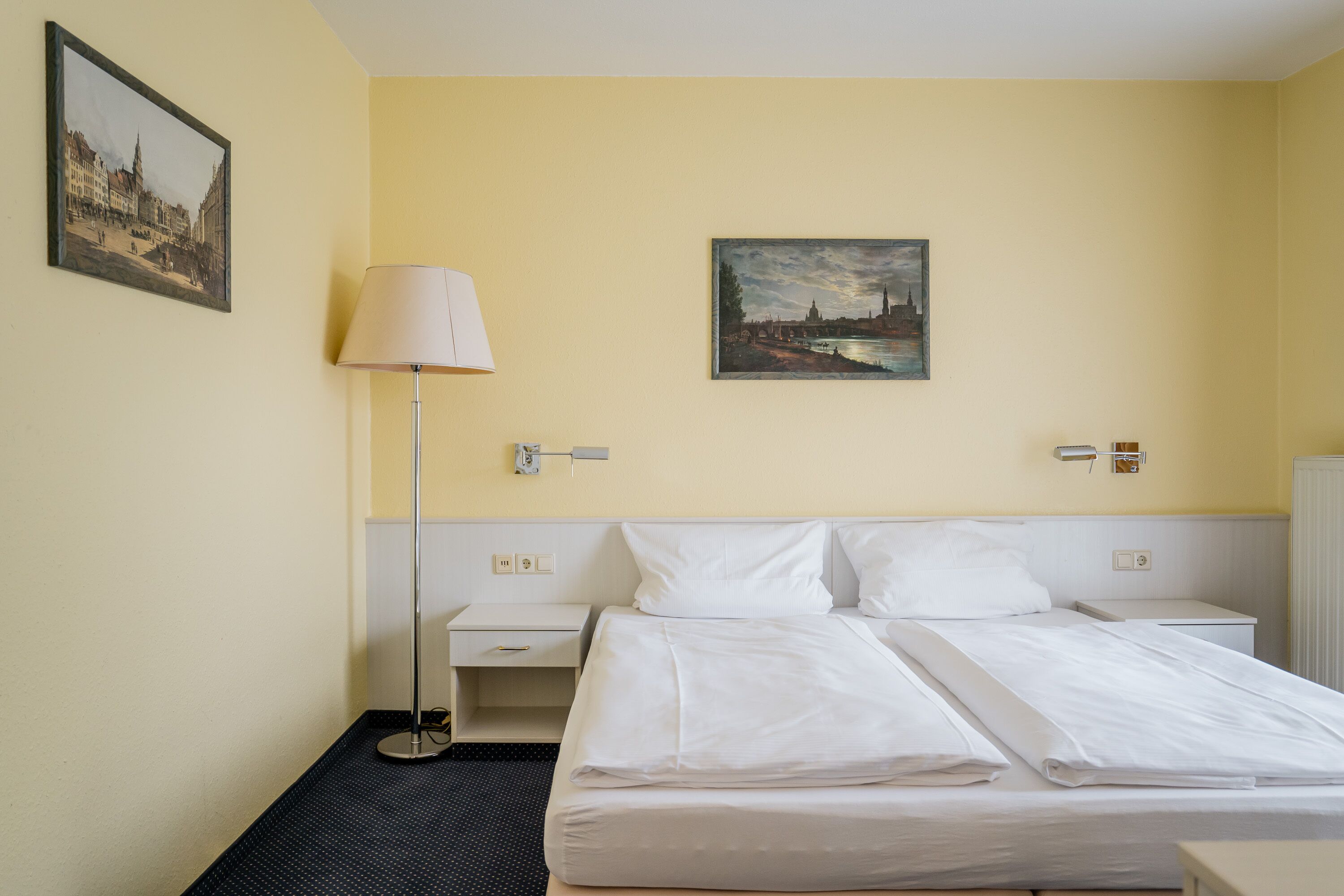 Ein Hotelzimmer mit Bett, Nachttisch, Lampe und zwei Bildern an der Wand.