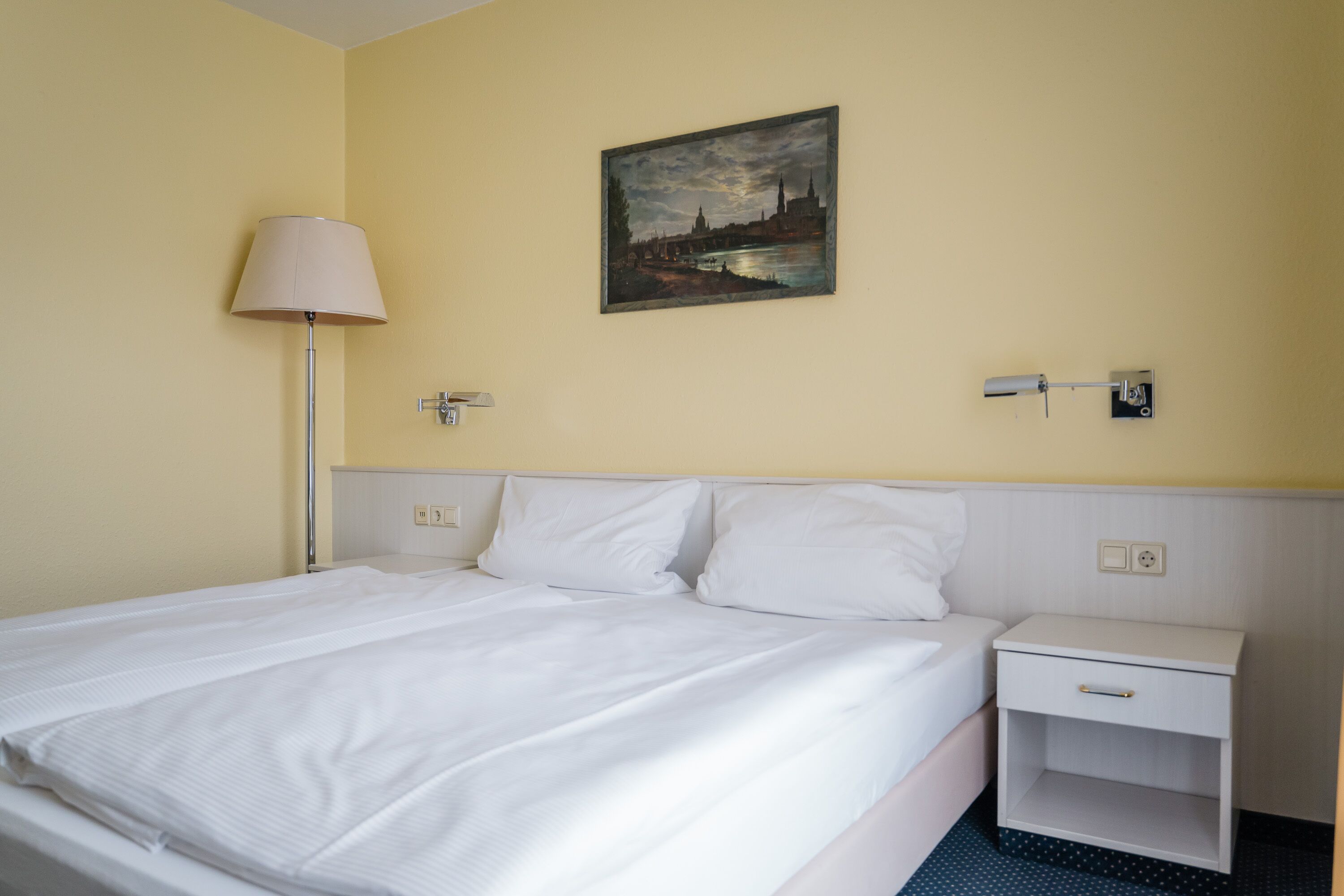 Een hotelkamer met een bed, nachtkastje, lamp en een schilderij aan de muur.