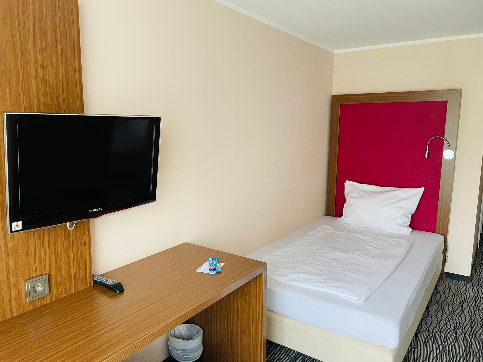 Una habitación de hotel con una cama, televisor, escritorio y un cesto de basura.