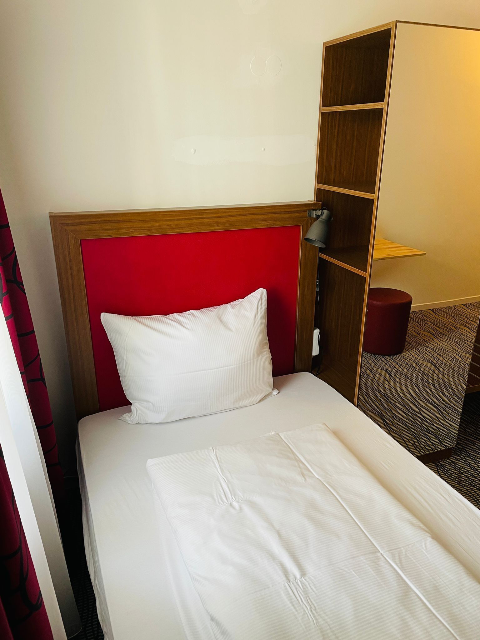 A hotel room with a bed, a red headboard, a wooden wardrobe, and a red stool.