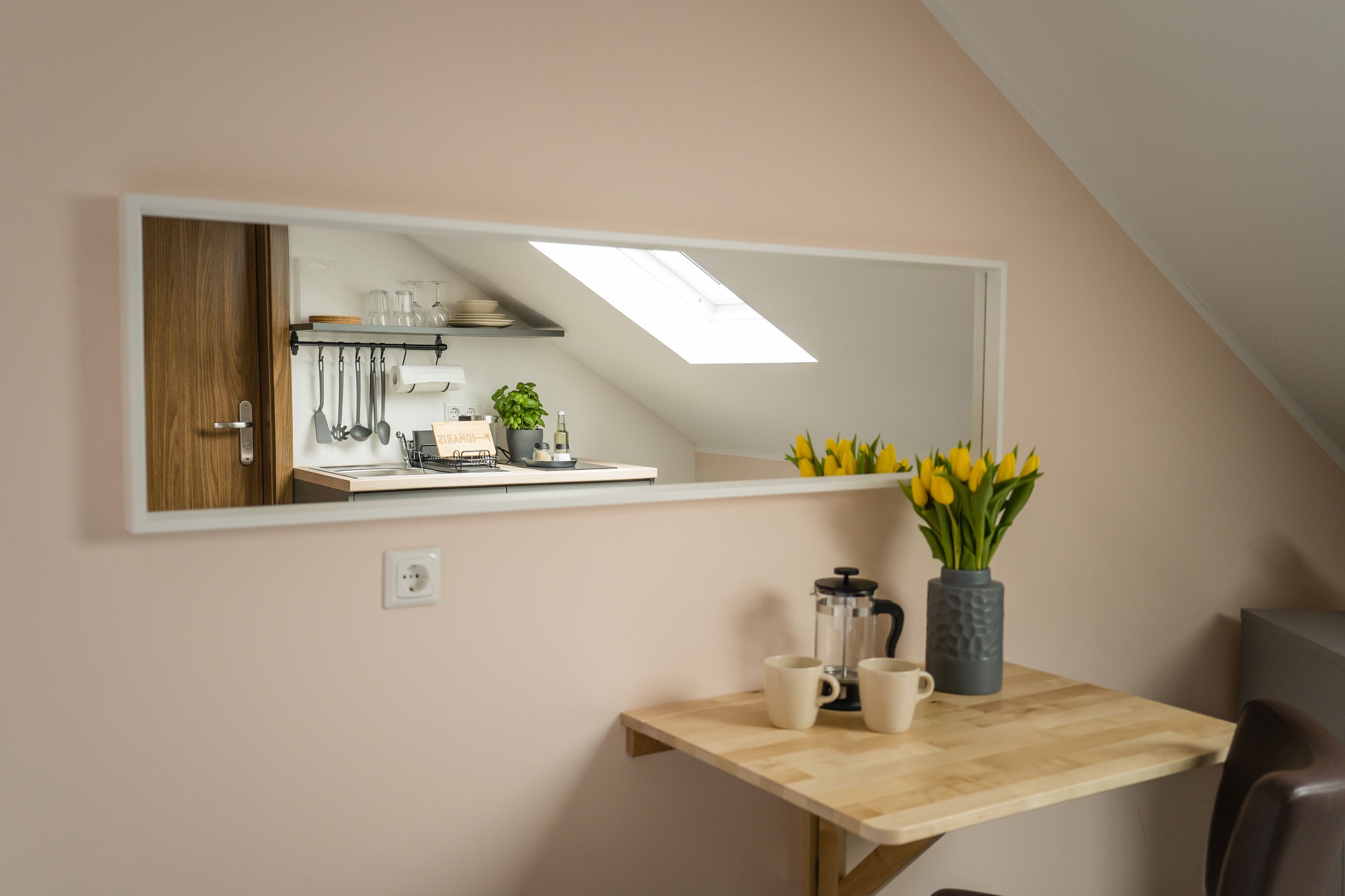 A kitchen with a large mirror, a wooden table with two cups, a coffee maker, and a vase with yellow flowers.