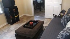A living room with a grey sofa, patterned cushions, a brown ottoman, and a black coffee table with a red object. - Lovely Midtown Home Close To ALL Downtown Action (New Orleans)