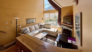 Un salon avec un canapé, une table basse et un téléviseur. Il y a une fenêtre avec des rideaux et une lampe.