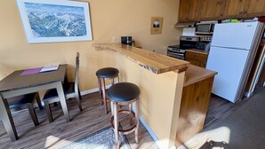 Une cuisine compacte avec des meubles en bois, un petit îlot central, une table à manger avec deux tabourets et un tableau encadré au mur.