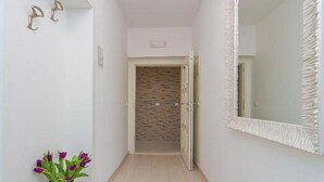 Un pasillo estrecho con piso de baldosas blancas, una puerta que da a un cuarto con pared texturizada y un espejo grande con un marco decorativo en la pared derecha.