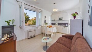 Una sala de estar moderna con un sofá marrón, un televisor y una zona de comedor con una mesa y sillas. Se observa una cocina al fondo con armarios blancos y un refrigerador.
