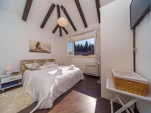 Relax in a serene bedroom with a large bed, nightstand, and bedside table, featuring a beautiful painting and a window with a view of the surrounding trees.