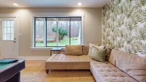 Una sala de estar con un sofá color beige, una mesa de billar y un ventanal con vistas a un jardín.