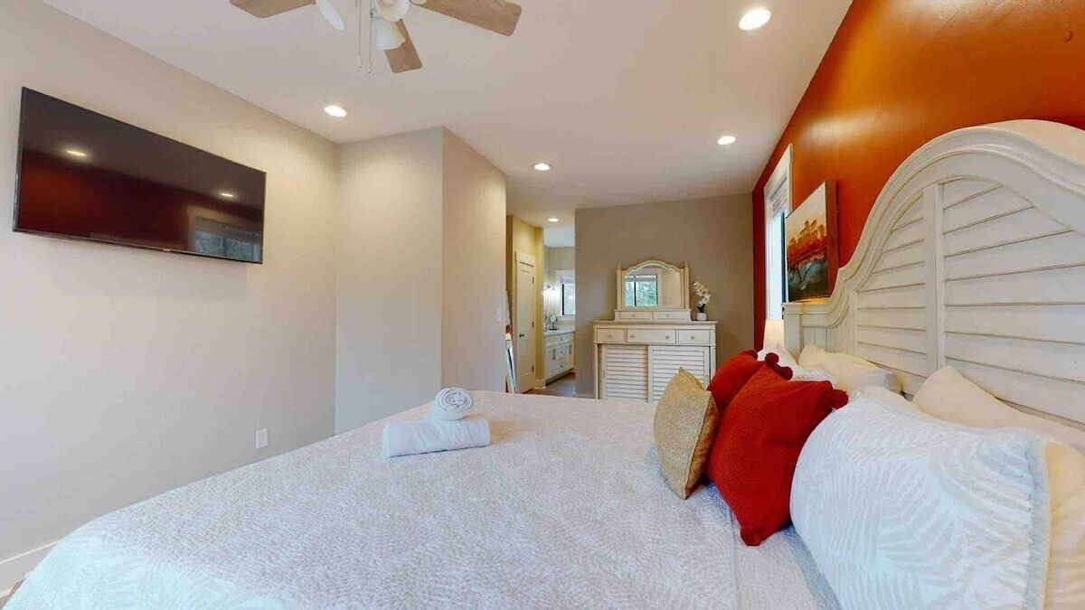 A bedroom with a bed, a ceiling fan, a TV mounted on the wall, and a dresser with a mirror.