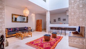 A spacious living area with a sofa, coffee table, and a vibrant rug. A bar area with stools is visible in the background. - Riad Bensaleh 56 (Marrakech)