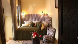 A bedroom with a patterned headboard, a bed with multiple pillows, a chair, a small table with a flower arrangement, and a patterned rug. - Riad Bensaleh 56 (Marrakech)