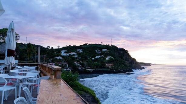 A coastal terrace with white chairs and tables, overlooking the sea and a hillside with buildings. - ATAMI- BEST PRIVATE BEACH HOUSE IN SURF CITY
walk to EL Tunco, El Zonte, Sunzal (La Libertad)