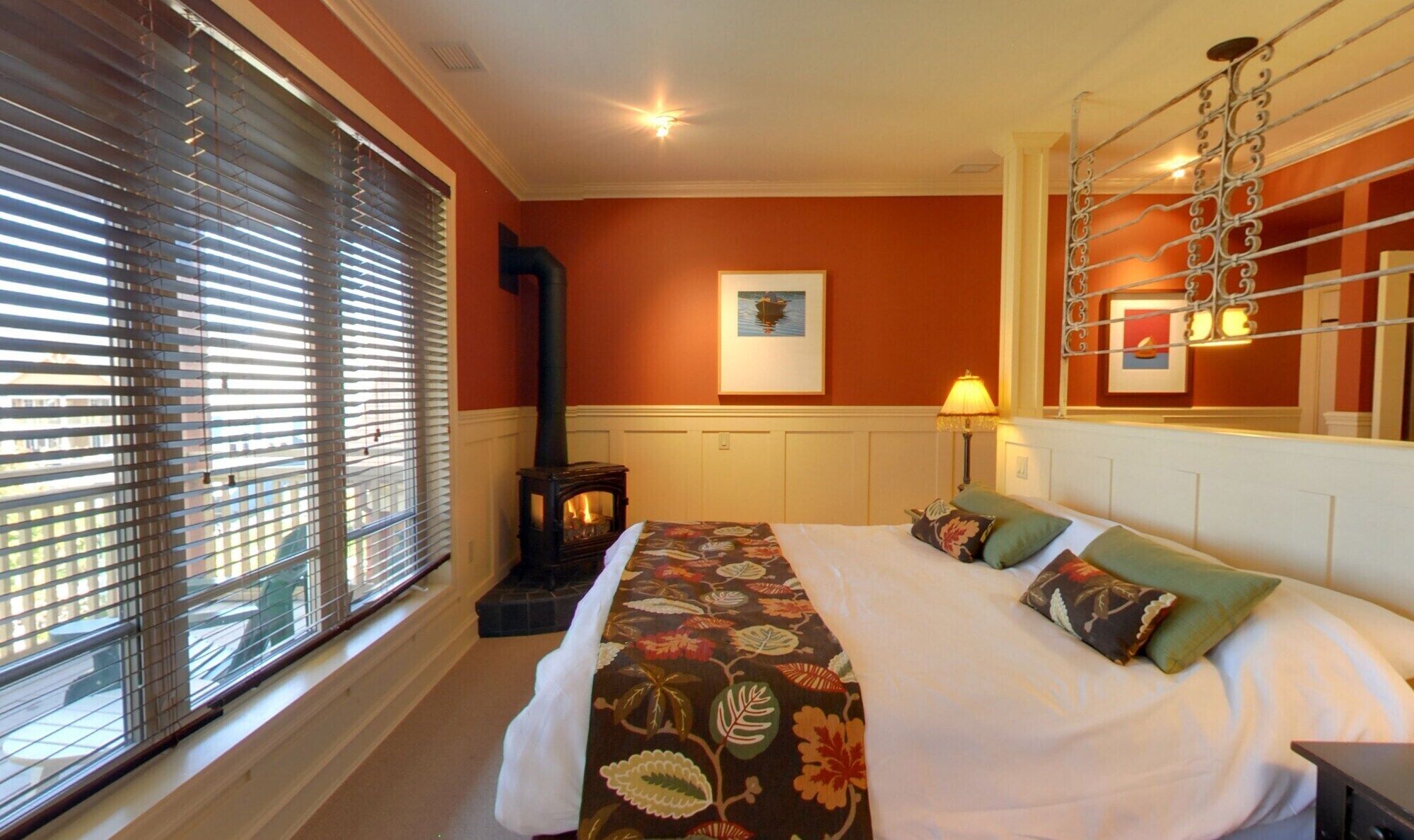 A hotel room with a bed, a fireplace, a window with blinds, and a painting on the wall.