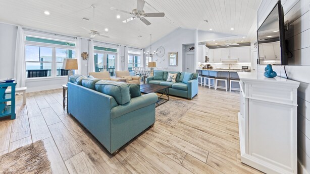 A spacious living room with a large blue sofa, a coffee table, and a ceiling fan. There is a kitchen area with white cabinets and a large window. - Early DISCOUNT heated pool 6Bed4Bath BeachVilla Steps2Sand (Gulf Shores)