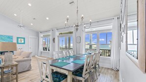 A dining area with a large table, white chairs, and a chandelier. There are windows with curtains, a ceiling fan, and a view of the ocean. - Early DISCOUNT heated pool 6Bed4Bath BeachVilla Steps2Sand (Gulf Shores)
