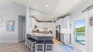 A modern kitchen with white cabinets, a central island with bar stools, a large window with a view of greenery, and a ceiling fan. - Early DISCOUNT heated pool 6Bed4Bath BeachVilla Steps2Sand (Gulf Shores)