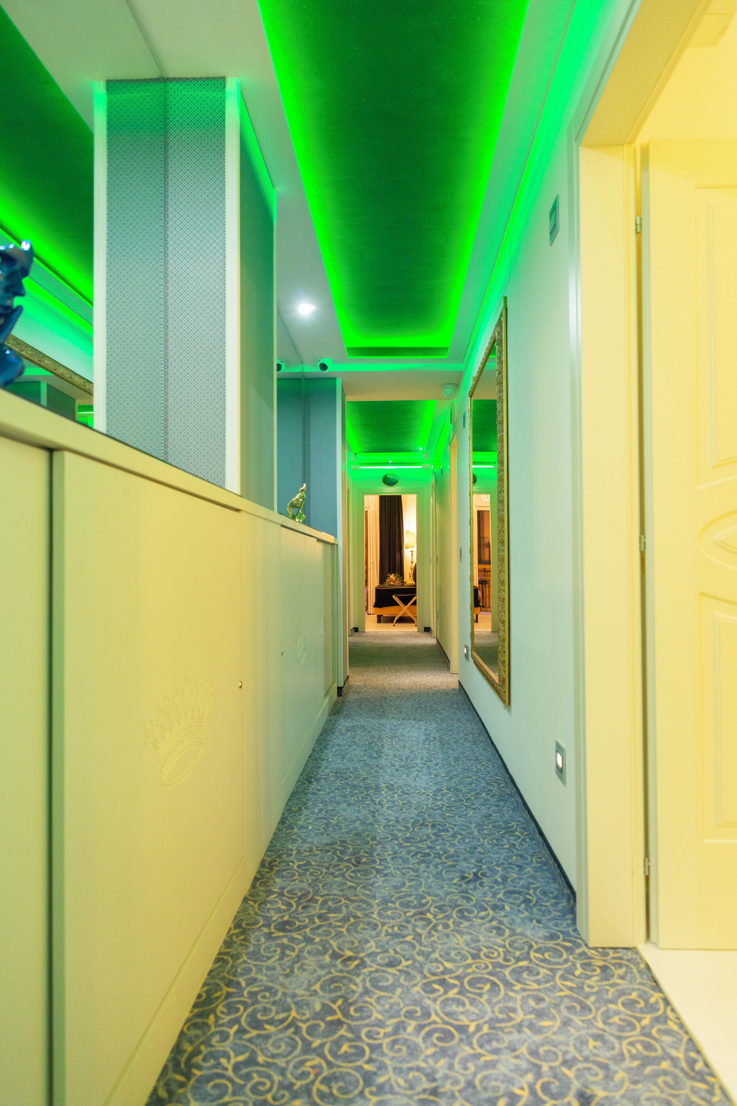 A narrow hallway with patterned carpet, green ceiling lights, and a door on the right.