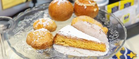 Una fuente de vidrio con varios pasteles, incluyendo una rebanada de pastel y rosquillas redondas, sobre una mesa con mantel estampado.