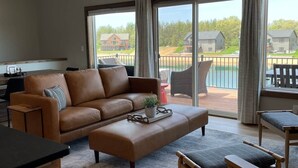 A spacious living area with a large window, a brown leather sofa, a wooden coffee table, and a view of a residential area. - Kokomo Cabin: Cozy Fires, Family Fun & Lake Views (Germantown)