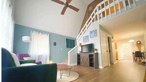 Una sala de estar con un sillón verde, una mesa de centro y un televisor sobre un mueble de madera. Una escalera con barandilla blanca conduce a un segundo piso.