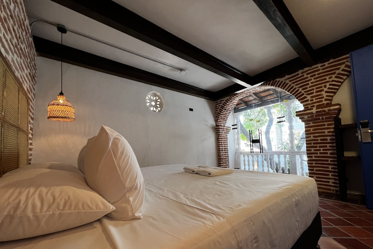 A bedroom with a bed, pillows, a hanging light fixture, a decorative wall piece, and a view through an arched window.