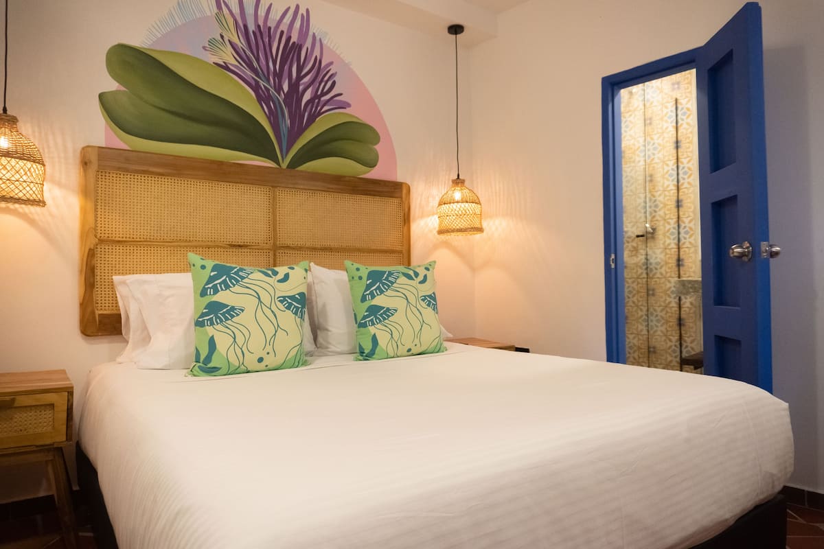 A neatly made bed with a white comforter and green patterned pillows, a woven headboard, and a blue door with a patterned glass panel.