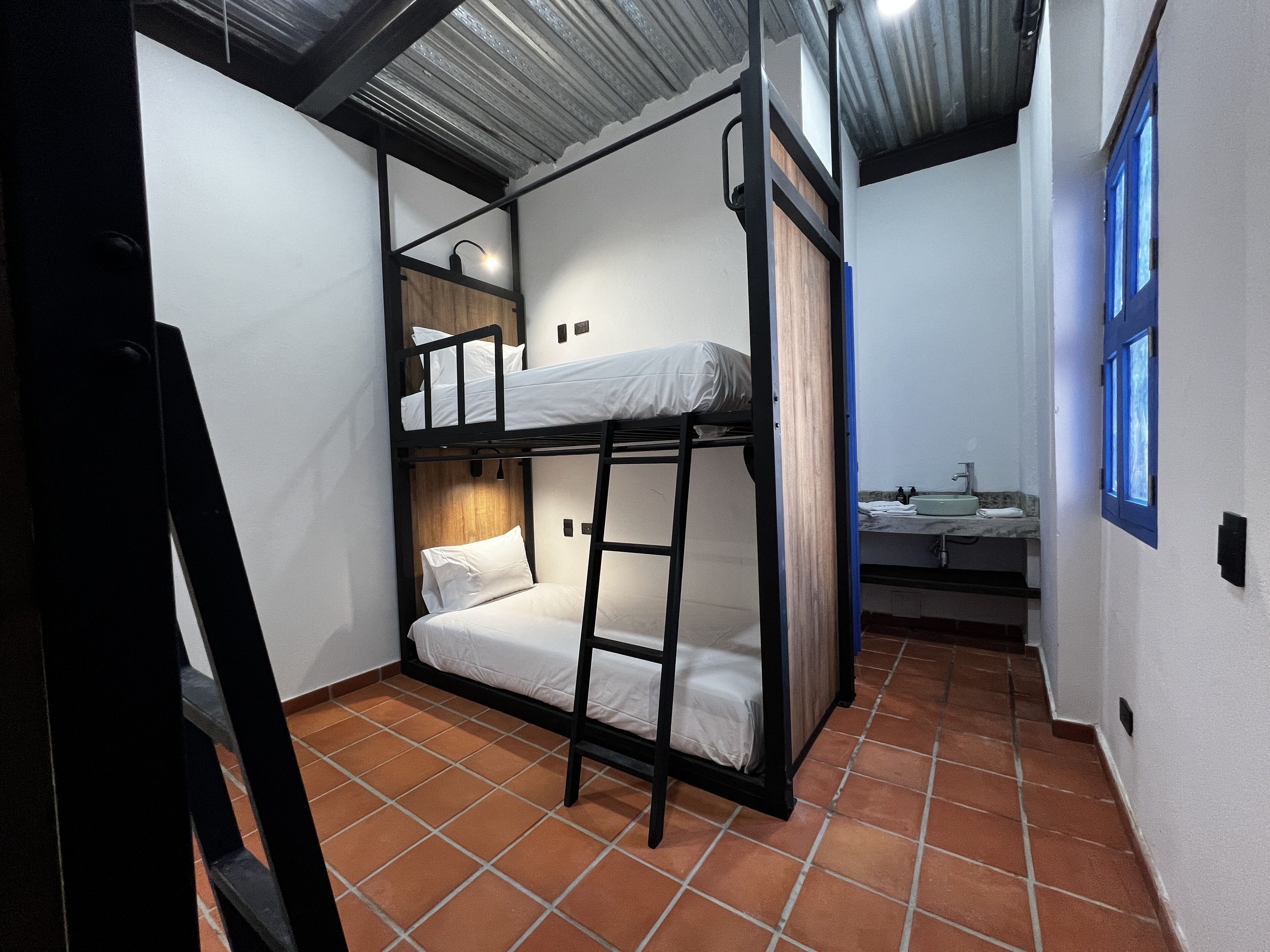 A compact room with a bunk bed, a small bathroom with a sink, and a window with blue shutters.