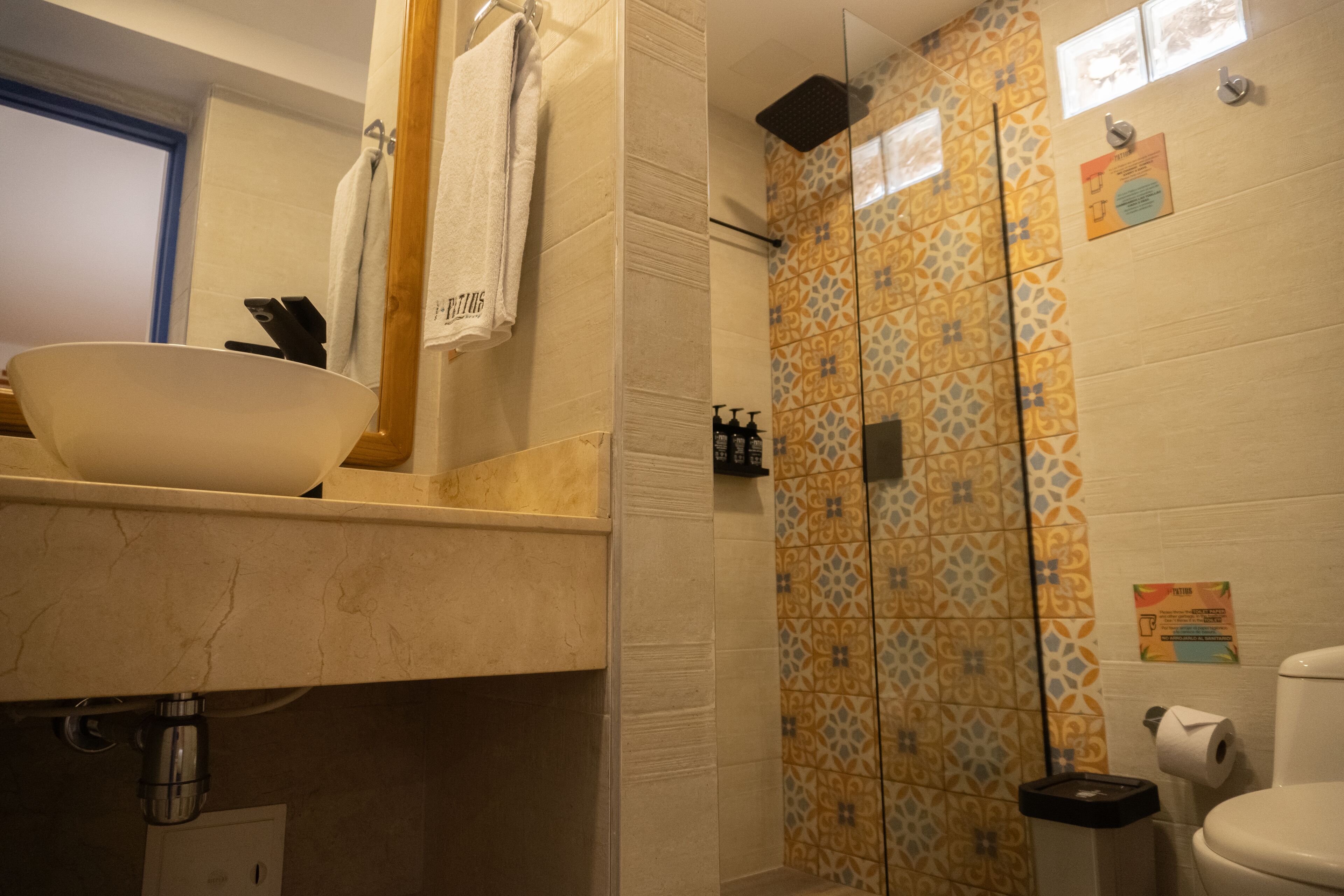 A bathroom with a sink, mirror, towel rack, shower with patterned tiles, toilet, and a trash bin.
