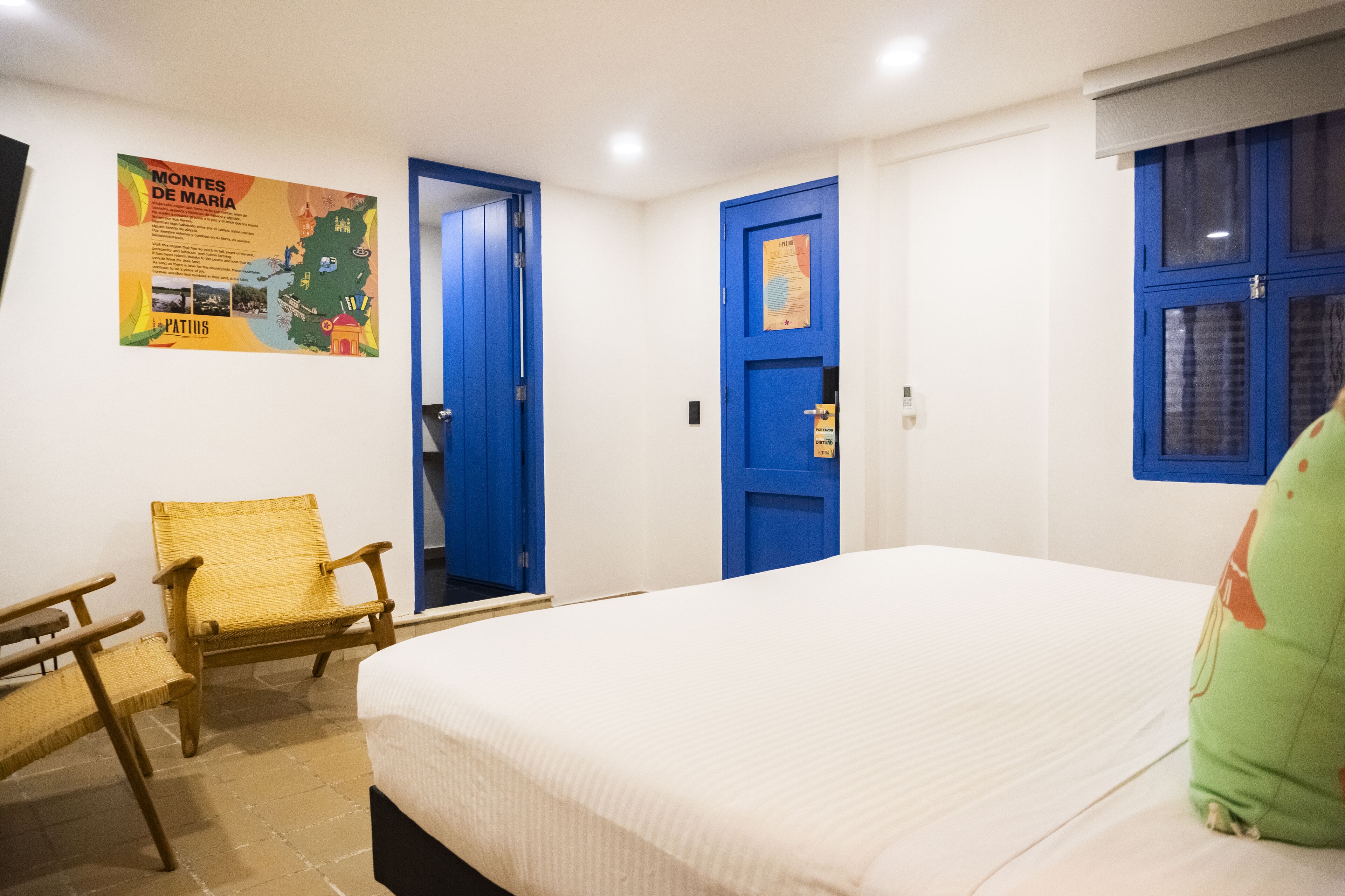 A hotel room with a bed, a chair, a blue door, and a poster on the wall.